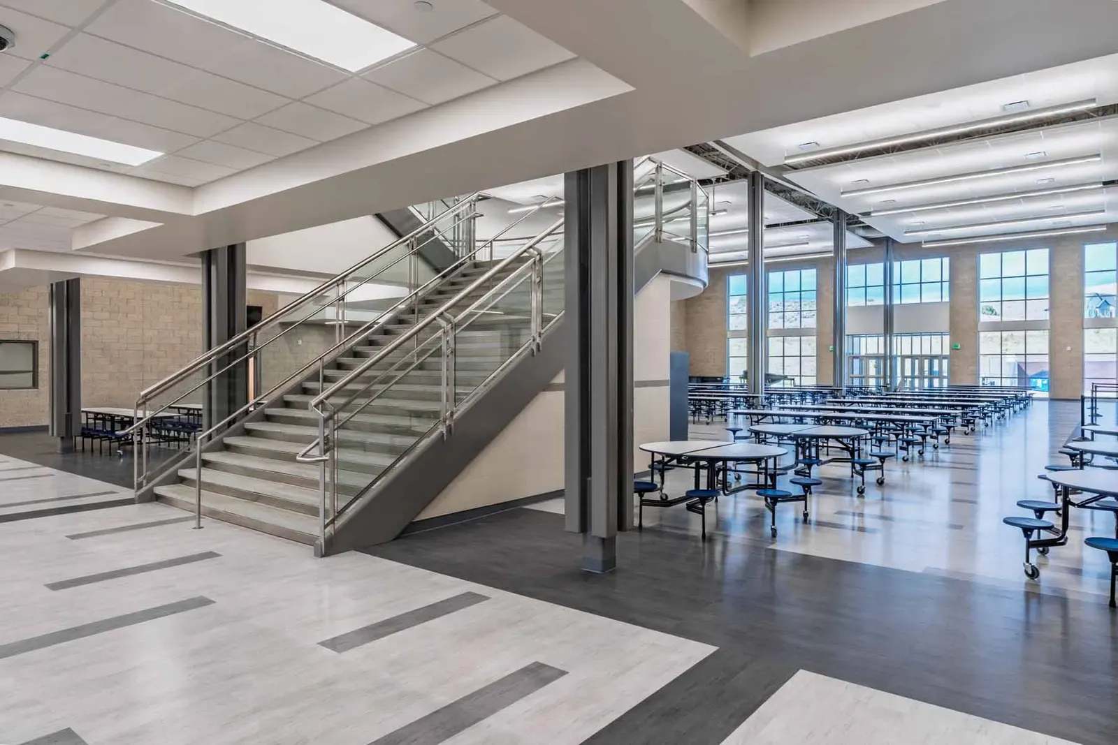 Commons with a glass-railed staircase, cafeteria tables and tall windows.