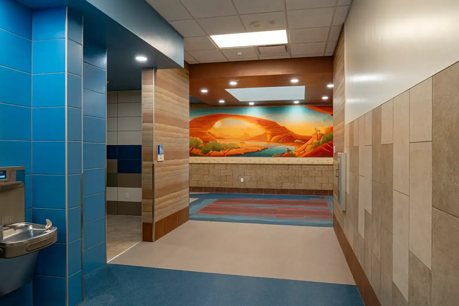 Corridor with blue tile walls, a drinking fountain and a desert landscape mural at the far end.