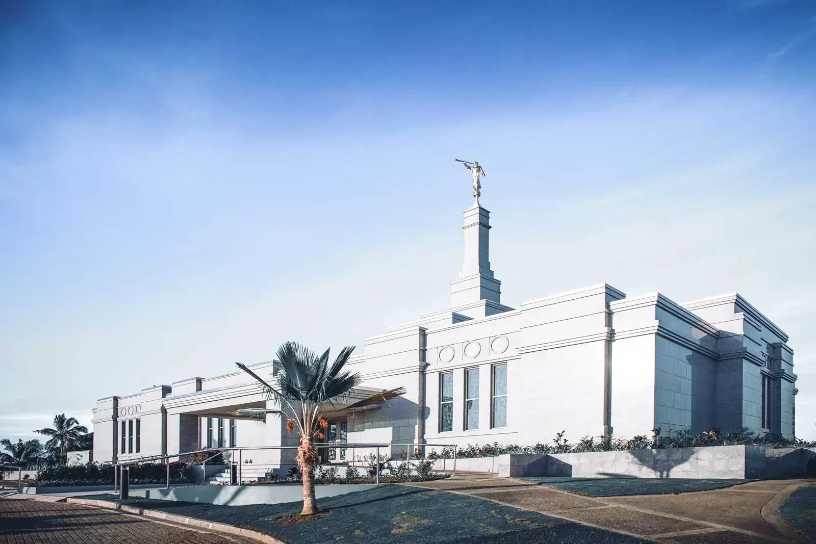 Side view of the temple's pale walls and spire topped with the Moroni statue, with palm trees in front under a blue sky.