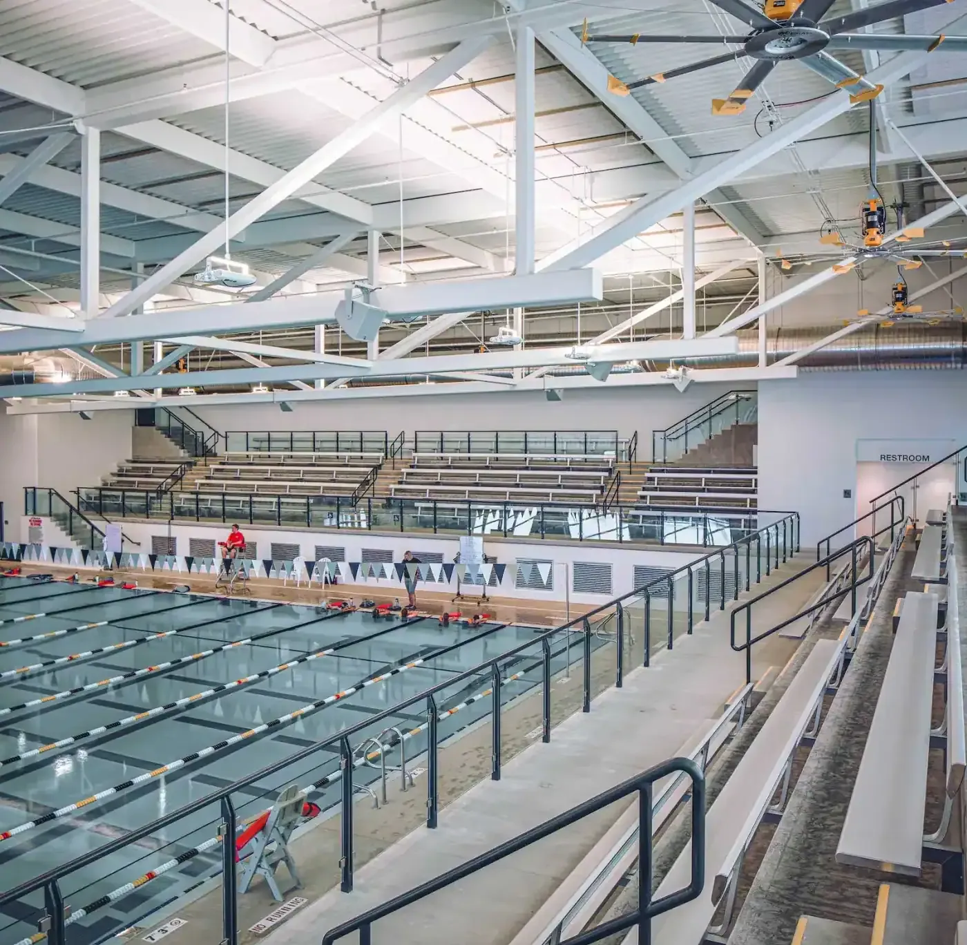 Elevated view of the competition pool from the spectator bleachers, with steel trusses overhead.