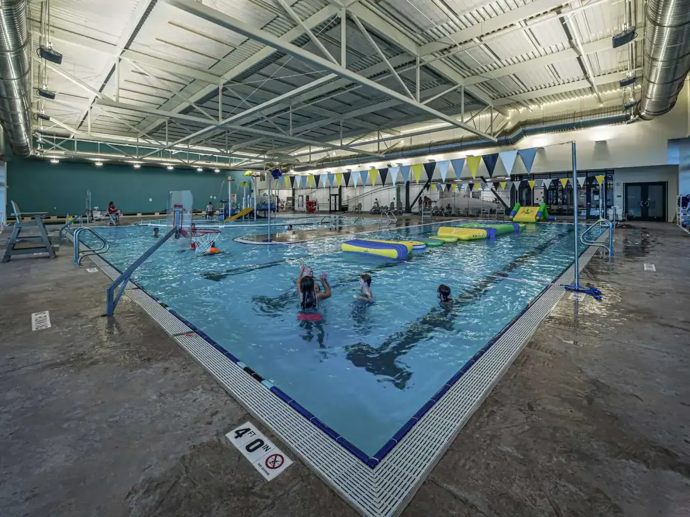 Pool with swimmers and a floating inflatable obstacle course beneath a steel truss roof.
