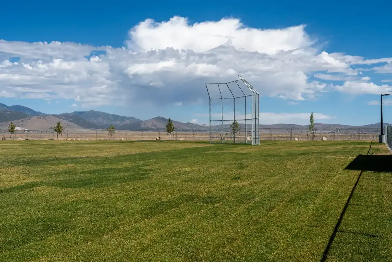 Grass field with a backstop and mountains under a cloudy blue sky.