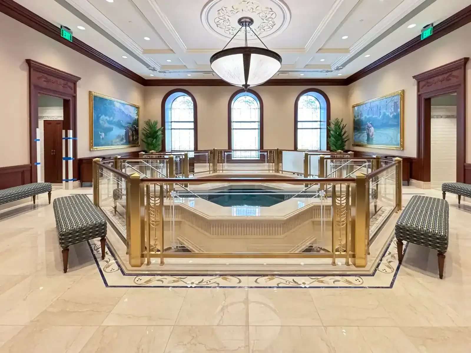 Baptistry with a font behind a glass and brass railing, arched windows, two large paintings and upholstered benches