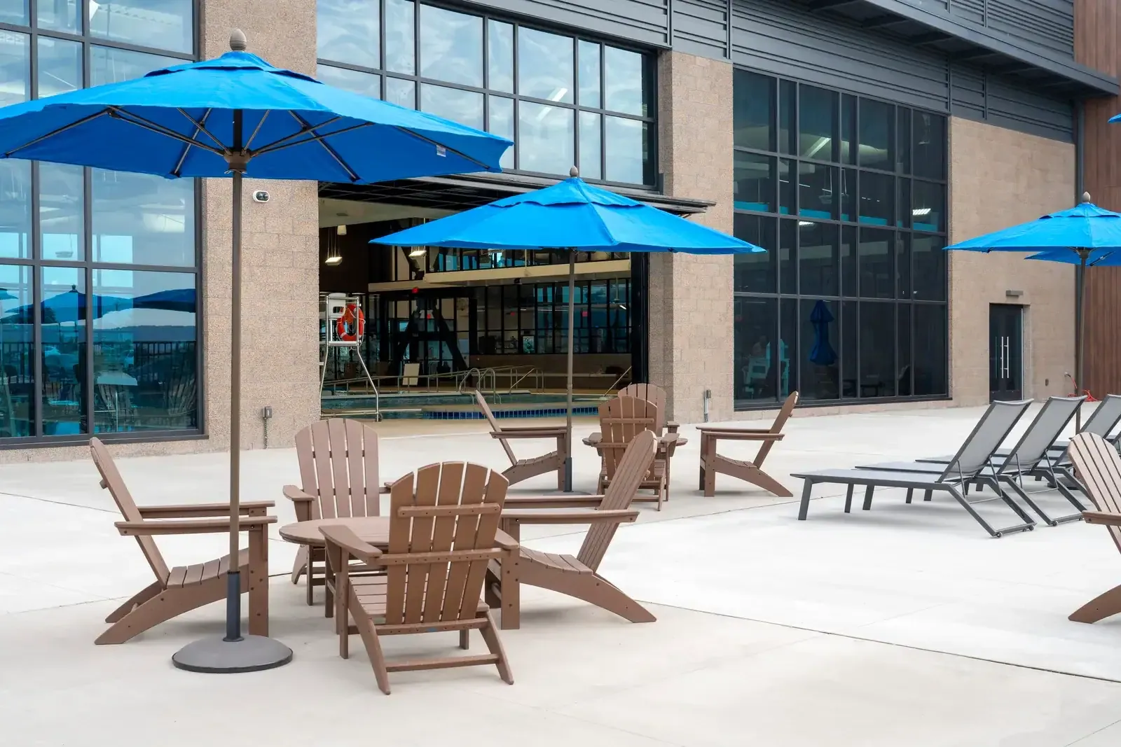 Outdoor patio with blue umbrellas, wood-look chairs and lounge chairs beside the building's glass wall and open pool door.