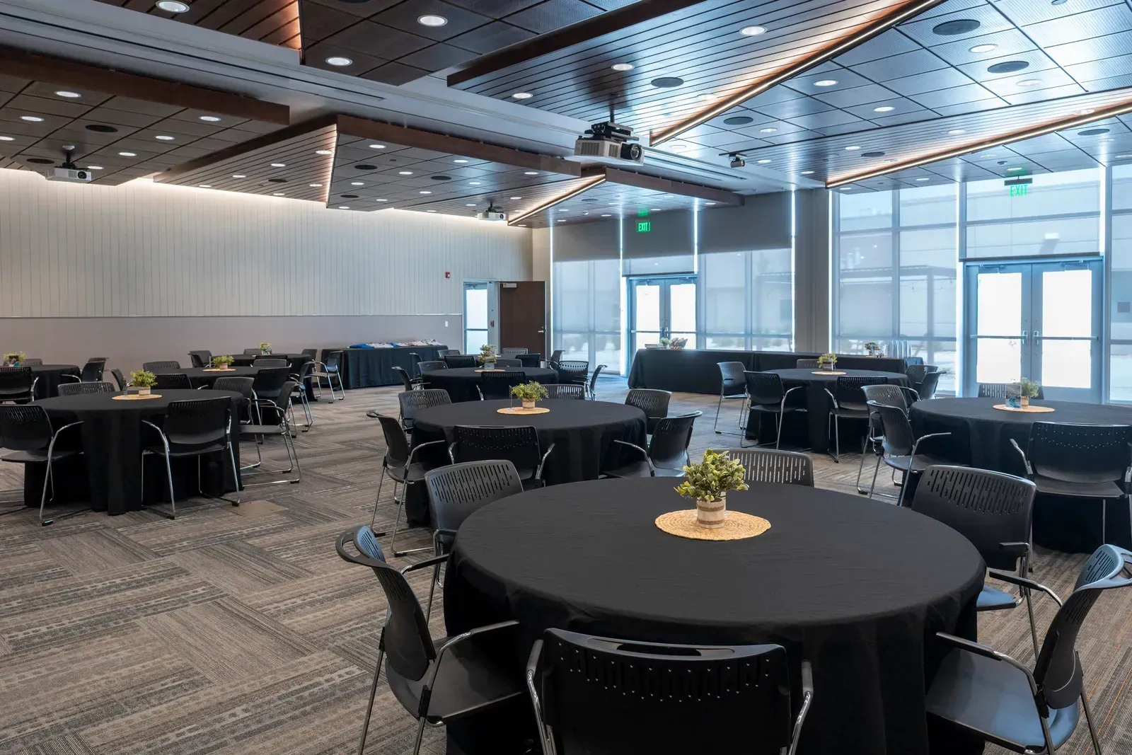 Community room set with round tables covered in black cloths, beside a wall of windows.