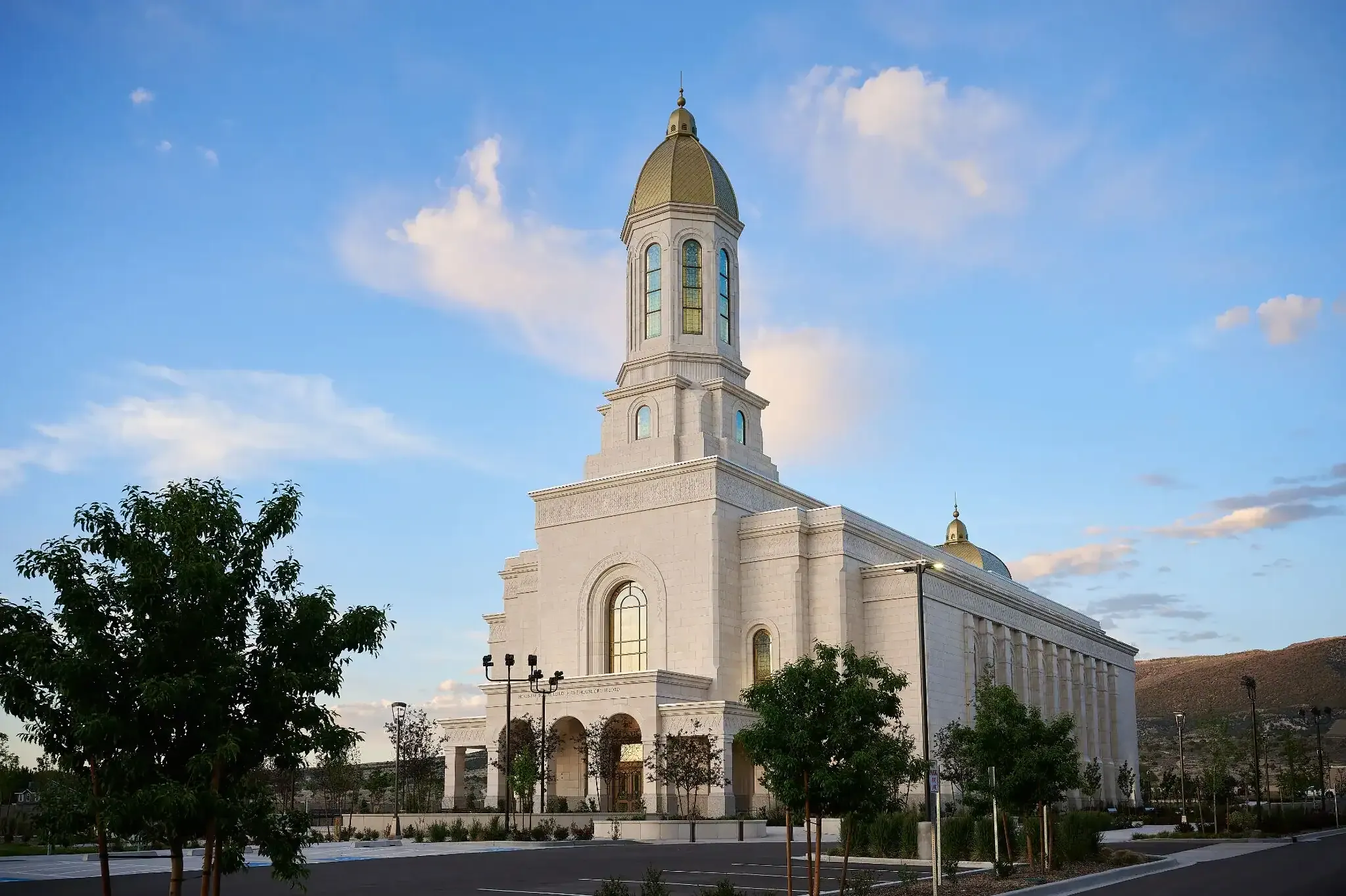 Exterior of the white temple with a gold-domed central tower against a blue sky, with young trees in the foreground
