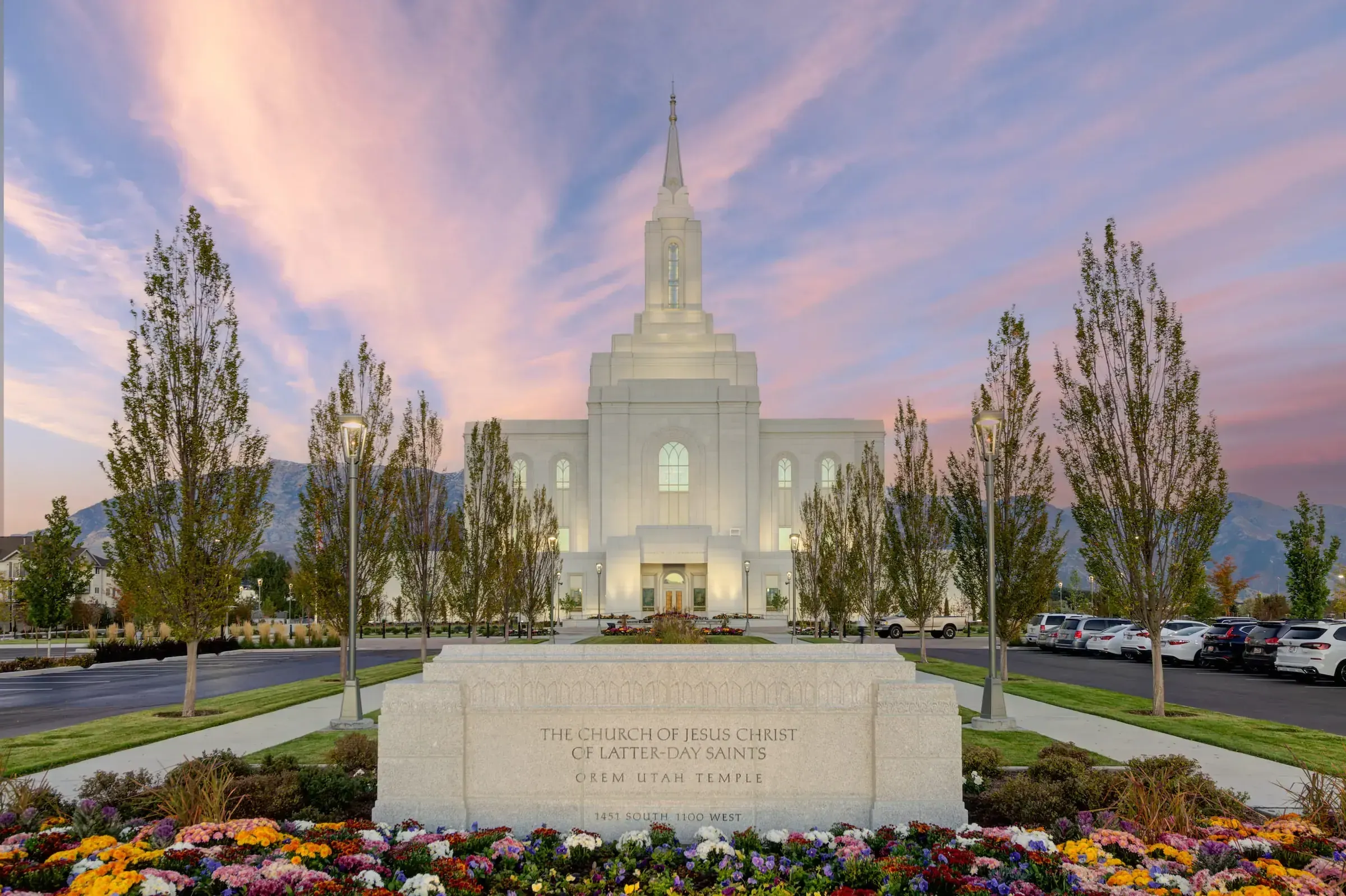 Orem Utah Temple at dusk behind its name sign and flower beds, with tall trees and mountains in the background.