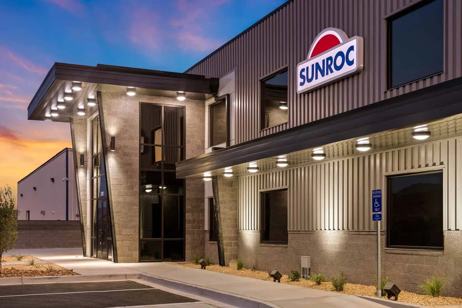 Office entrance at dusk with a Sunroc sign on gray metal siding, a stone-clad tower and lit canopies.