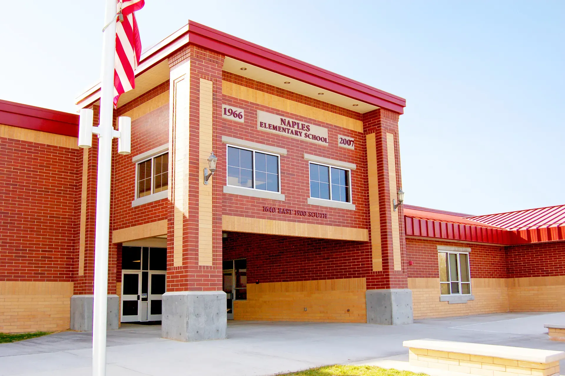 Brick entrance of Naples Elementary School with a red metal roof, the school name on the facade and a flagpole in front.