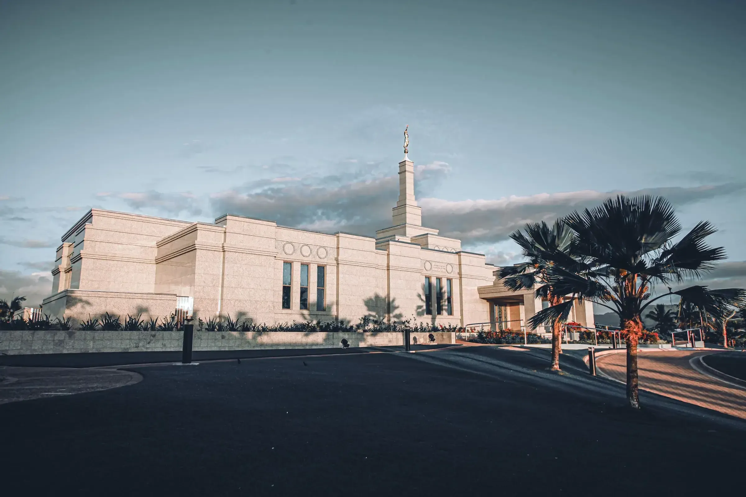 Exterior of the Fiji Temple with its tall central spire and palm trees beside the entrance drive under a cloudy sky.