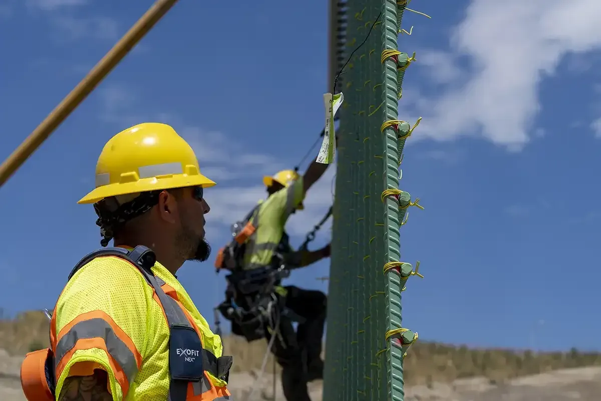 An ironworker in a yellow hard hat and harness beside a rebar column, with a second worker tied off above